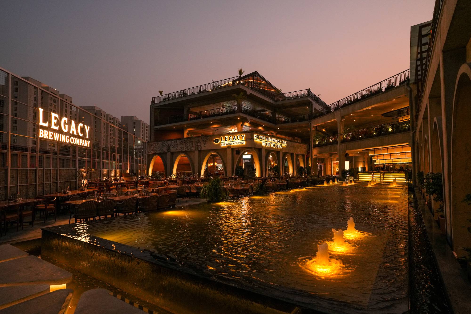 Legacy Brewing Company's glowing fountain plaza at dusk, framed by three floors of open-air dining